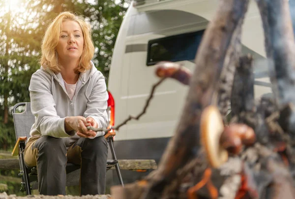 A lady grilling a hotdog on the camping site fire.